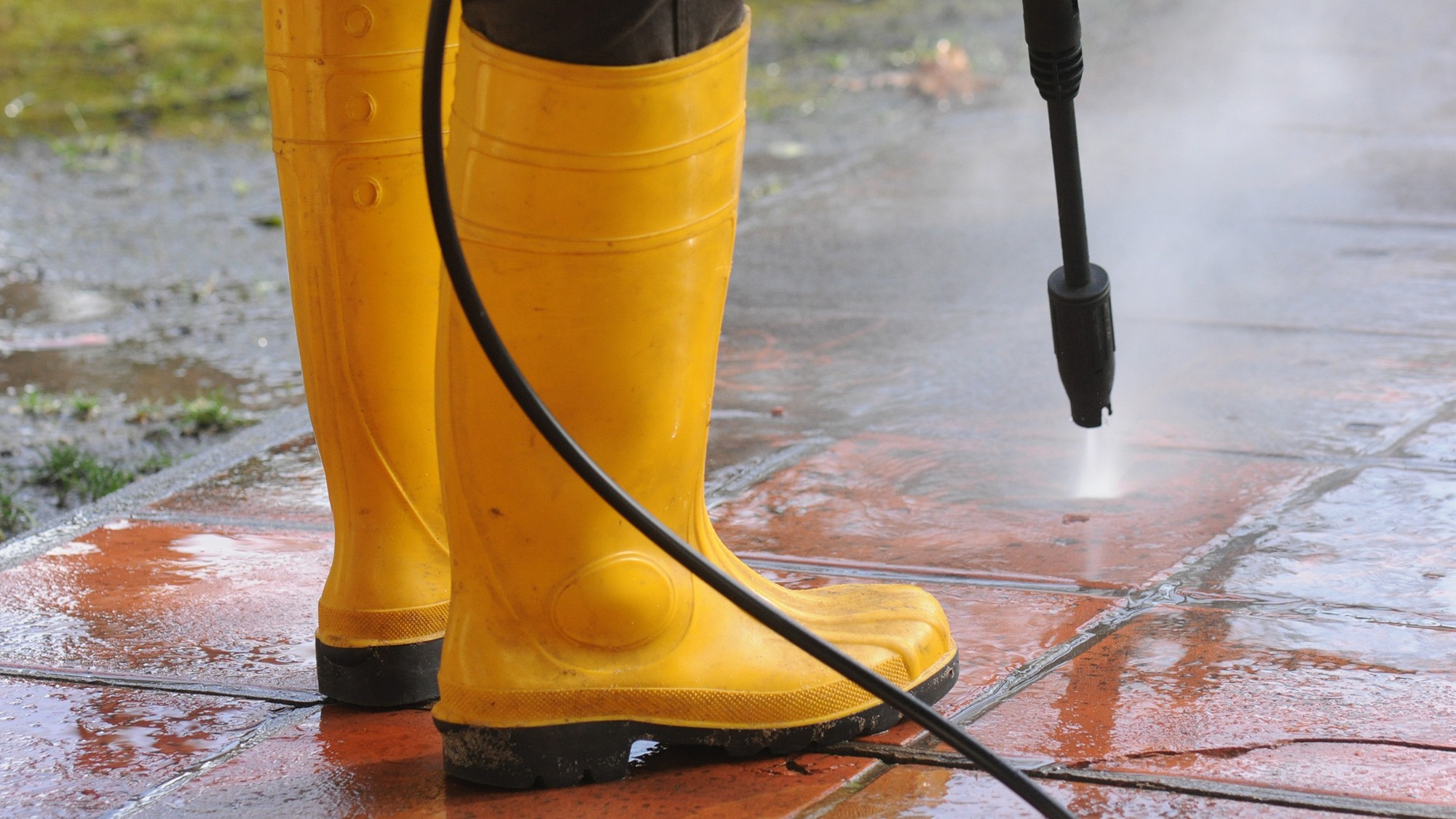 Person wearing yellow rubber boots with high-pressure water nozzle cleaning the dirt in the tiles